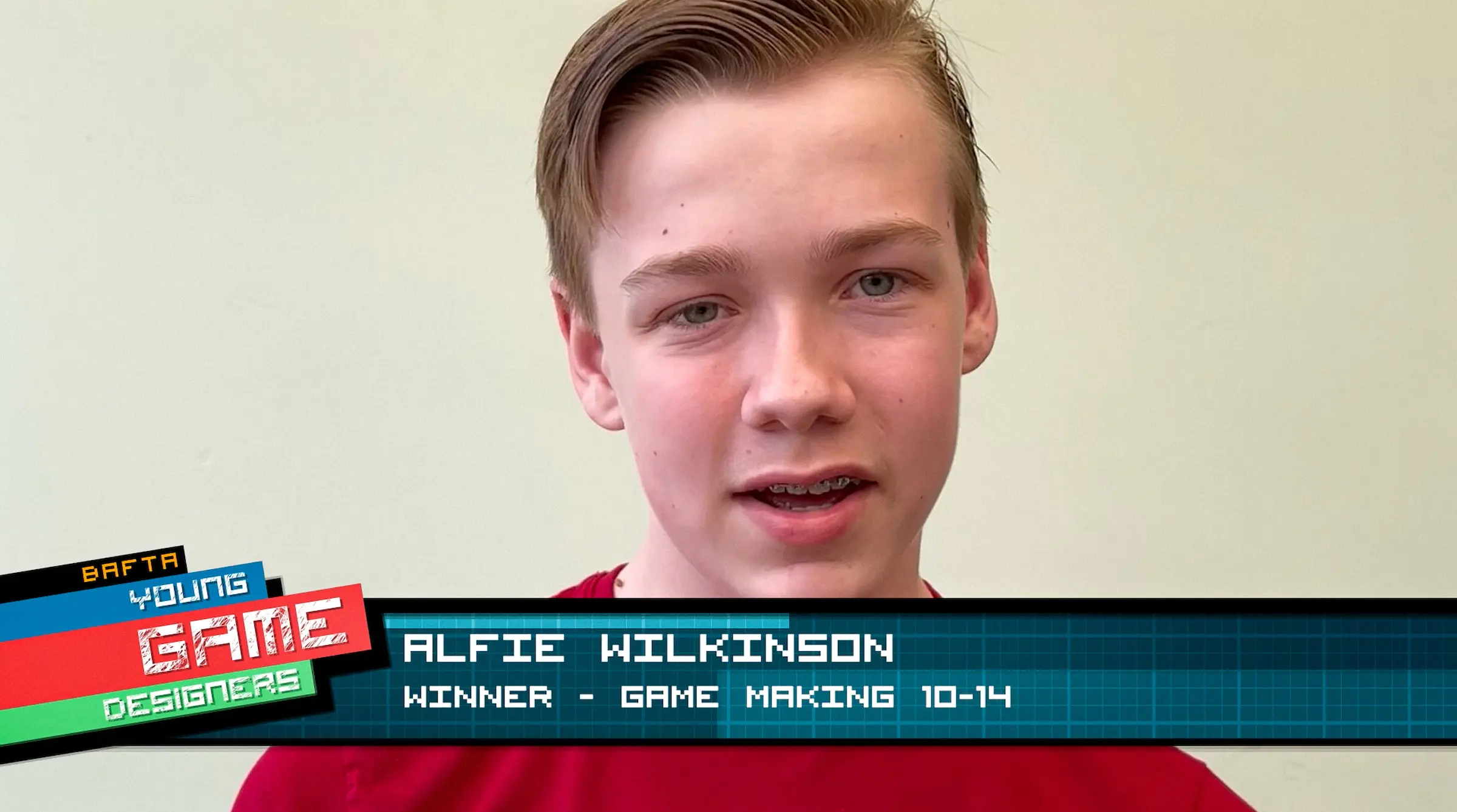 A young white boy with combed back blond hair and wearing a red t-shirt. There is a banner across the bottom of the screen that reads: BAFTA Young Game Designers - Alfie Wilkinson, Winner - Game Making 10-14.