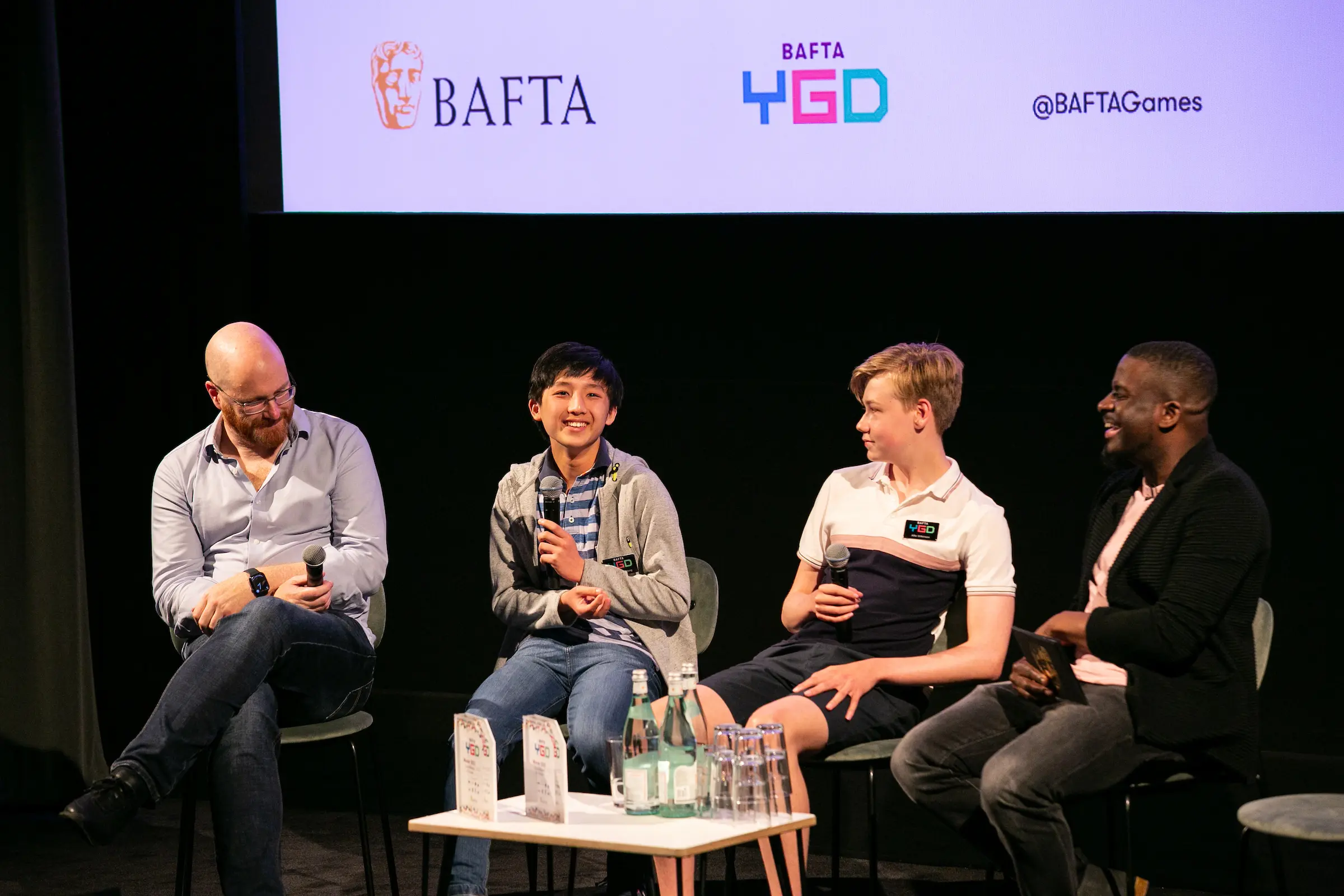 Four people sat on stage with microphones. From left to right there is a bald white man with glasses and a grey shirt, two boys - one Asian with dark hair and one white with blond hair, and a black man with closely shaved black hair and a black blazer.