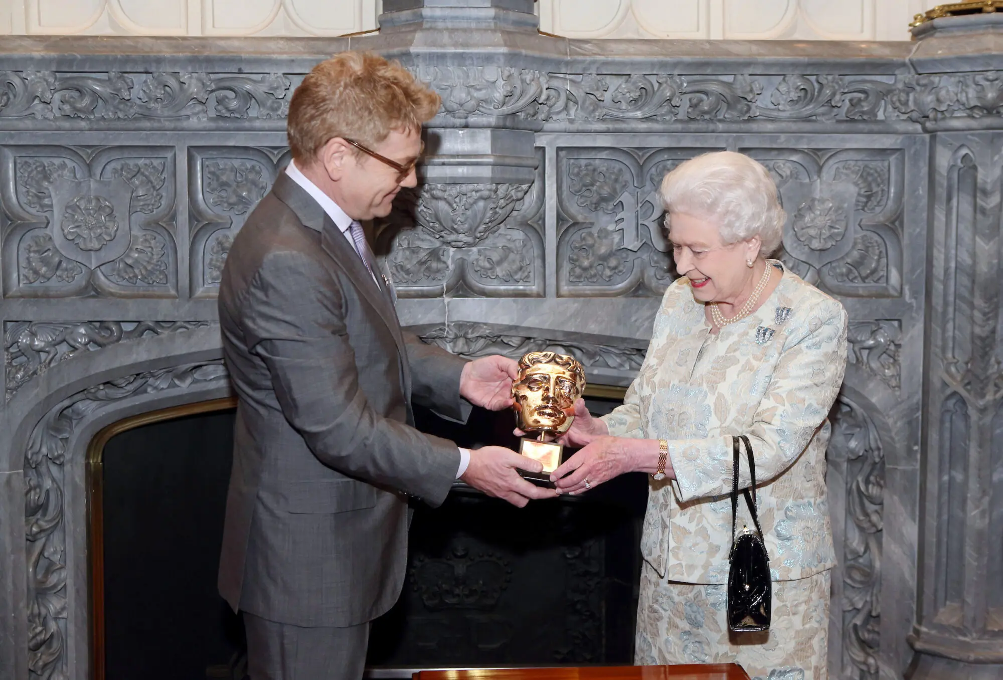 A man with short strawberry blond hair and glasses in a grey suit handing a BAFTA mask to the Queen, an older woman with white hair and a cream floral patterned suit.