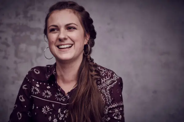 A woman with long brown hair the top of which is plaited. She is wearing a dark red shirt with a white pattern on.