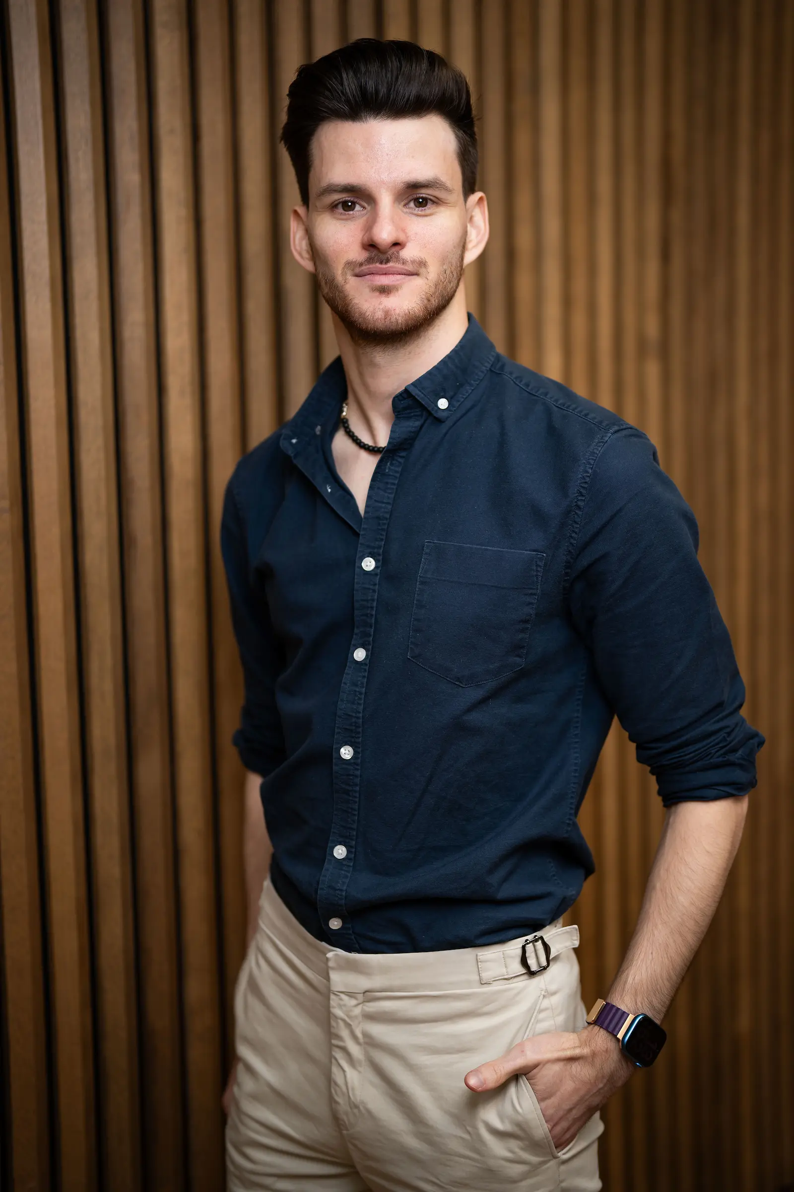 A white man with thick dark hair with short stubble. He is wearing a dark blue shirt with white buttons and cream trousers.