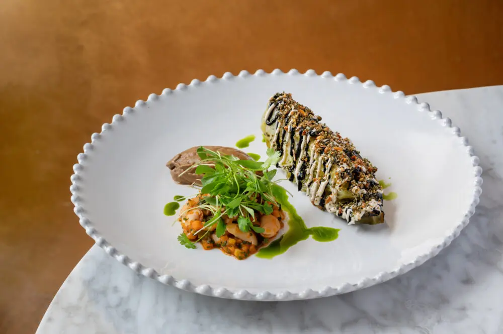 A wedge of chargrilled cabbage dressed with black and white sauces next to a quenelle of mousse and a portion of butter bean stew on a white plate.