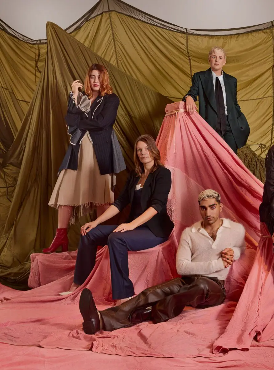 Four of the BAFTA 2024 Breakthrough cohort. On the left is a white woman with bobbed red hair wearing a denim blazer with statement sleeves. Second from left is a white woman with shoulder length light brown hair wearing a black top, black blazer and jeans. Second from right is an Asian man with short dyed blond hair sat on the floor wearing a white mohair top and brown leather trousers. On the far right stood at the back is a white woman with a short blonde pixie crop and wearing an oversized grey suit.