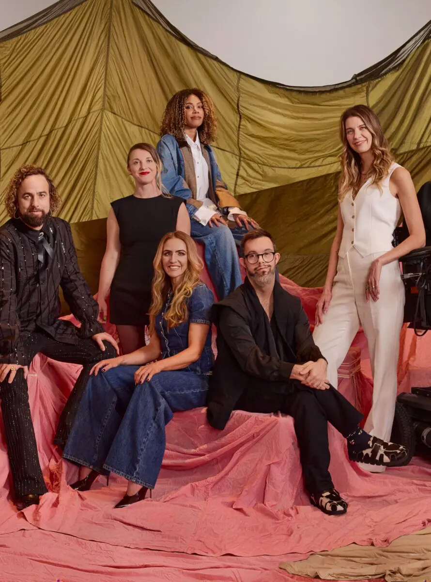 Six of the BAFTA 2024 Breakthrough cohort. On the left is a white man with short curly hair and a beard wearing a black shirt with sparkly stripe running through it. Next is a white woman with blonde hair tied up and bold red lipstick wearing a black shift dress. Next is a a white woman with long wavy blonde hair sat down wearing a denim jumpsuit. Sat at the back is a black woman with shoulder length tightly curled hair and wearing a white shirt and denim jacket. Second from right is a man with short dark hair, a grey beard, and black rimmed glasses sat down wearing a black suit. On the far right is a woman with long dark blonde slightly curled hair wearing a white waistcoat and trousers.