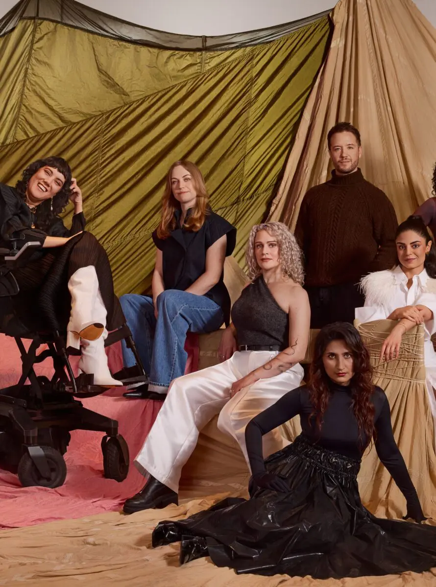 Six of the BAFTA 2024 Breakthrough cohort. On the far left is a white woman with short dark hair and a thick fringe, wearing an all black outfit, white boots and sat in an electric wheelchair. Next is a white woman with light brown shoulder length hair wearing a short sleeved black shirt and blue jeans. In the middle of the photo is a white woman with shoulder length curly white hair wearing a one shoulder black top and white trousers. On the right of the photo is a man with short dark hair and wearing a brown knitted turtleneck jumper stood at the back. Sat on the floor at the front of the photo is an Asian woman with long dark wavy hair wearing a black top and long black skirt. On the far right is an Iraqi woman with dark hair tied back wearing a white shirt with feathers on the shoulder.