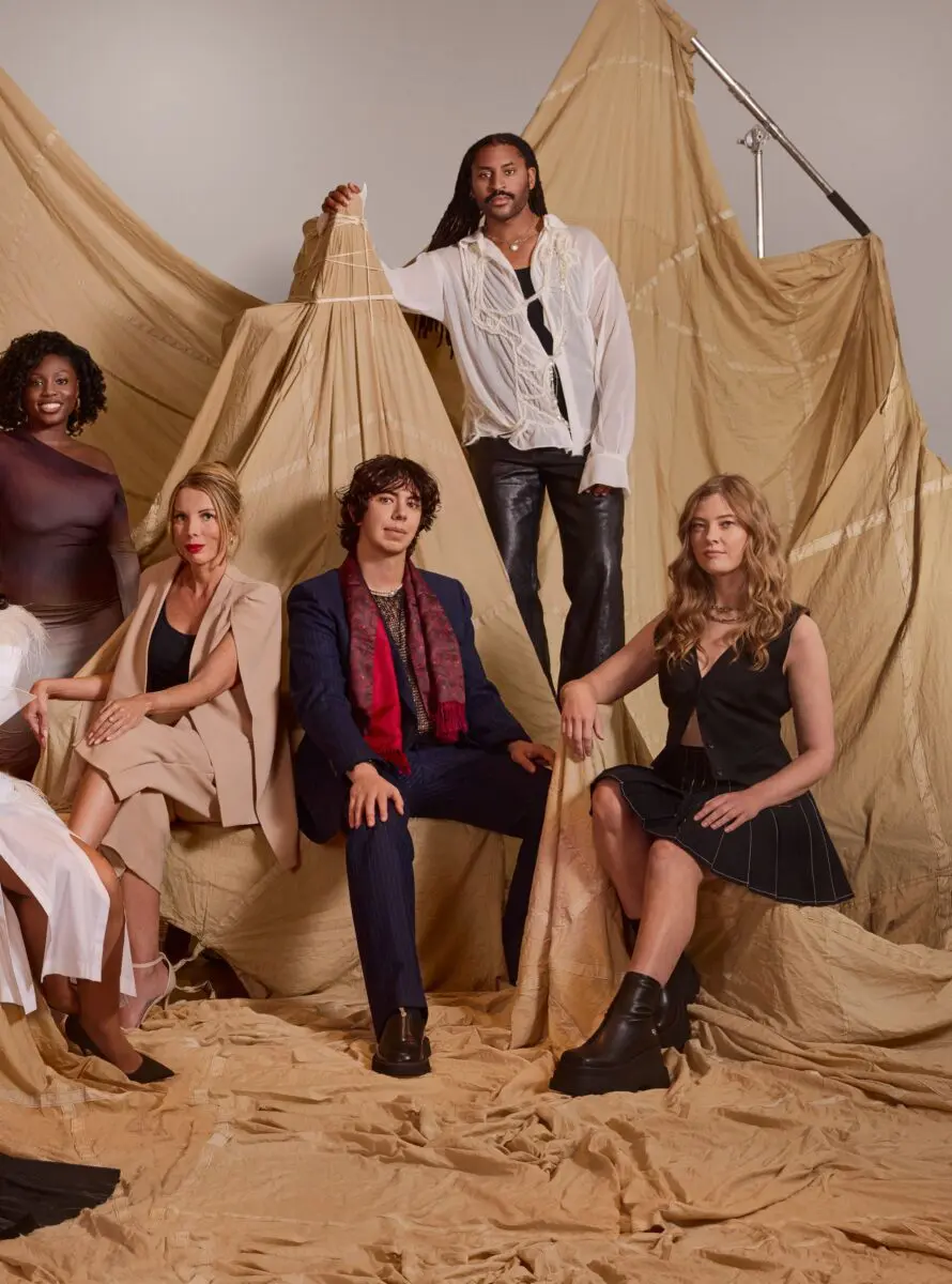 Five of the BAFTA 2024 Breakthrough cohort. On the far left is a black woman with short, dark, curly hair wearing a purple ombre one-shoulder dress. Next is a white woman with blonde hair tied up and bold red lipstick wearing a black top and pale pink caped suit. In the middle is a white man with short dark curly hair wearing a blue suit and a red patterned scarf. Next is a black man with long dark dreads wearing a black top, white floaty sleeved shirt and black leather trousers. On the far right is a white woman with long blonde curly hair wearing a black skirt with white stitching on and a black waistcoat.