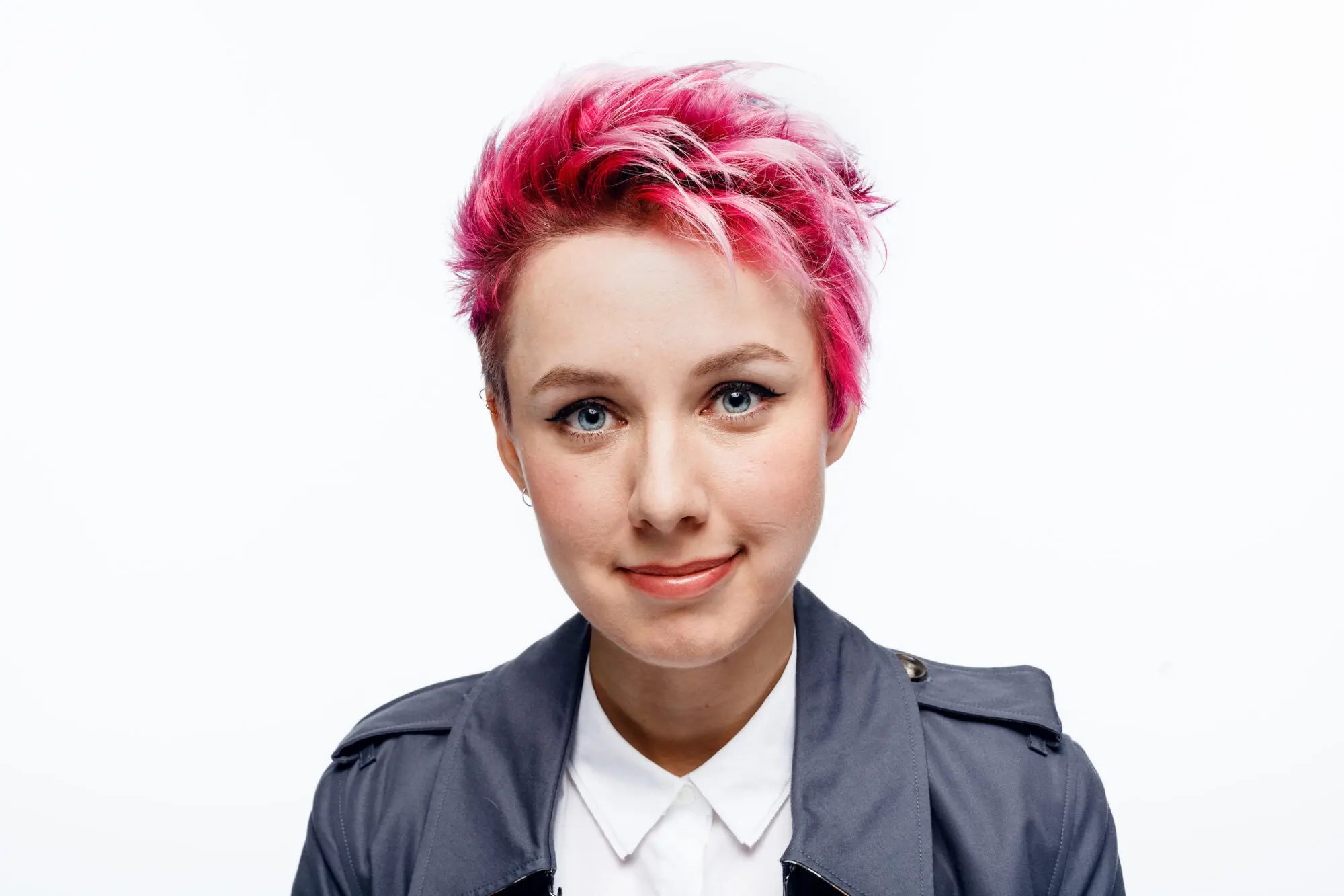 A woman with a dyed pink pixie crop wearing a white shirt and grey trench coat.