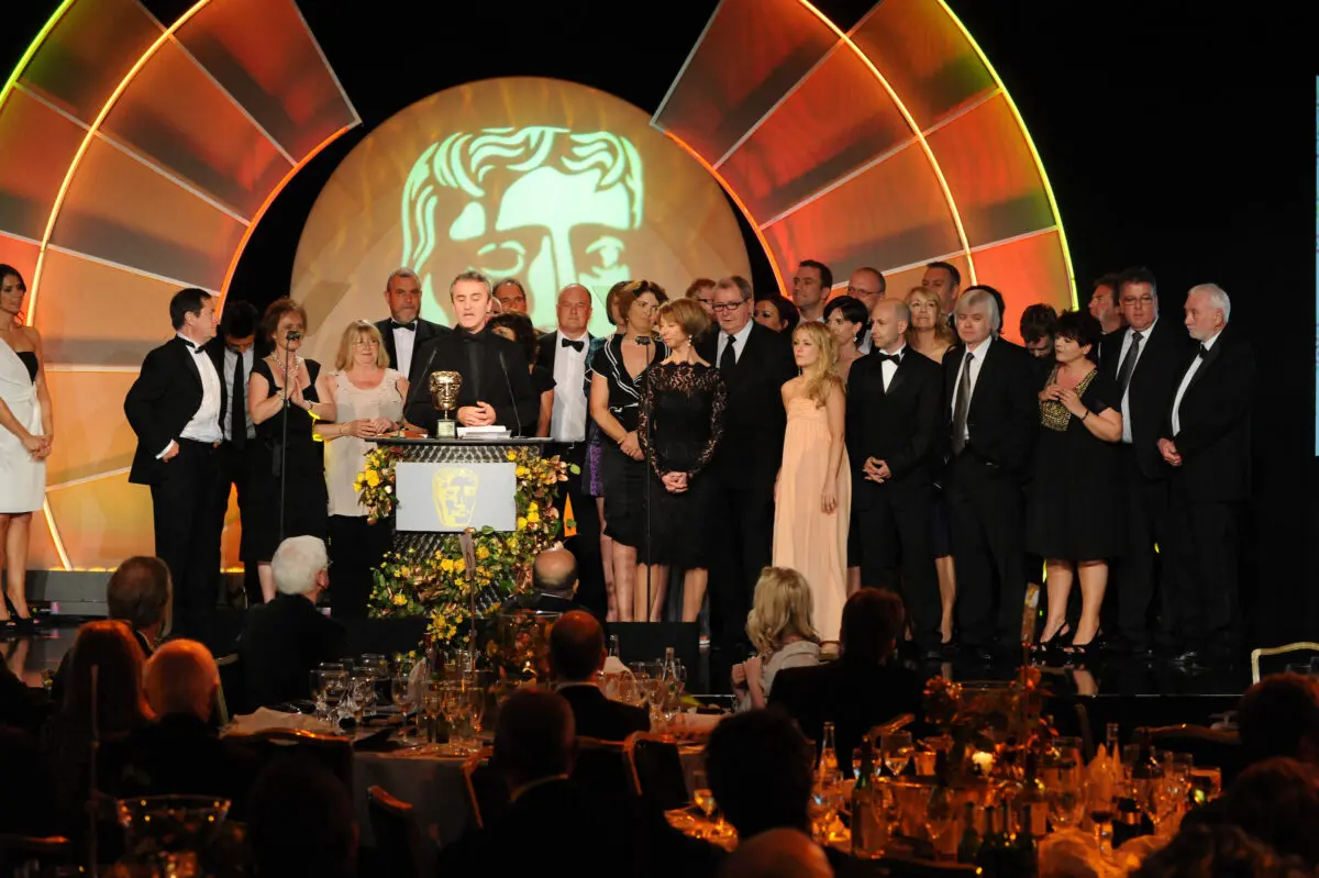A crowd of people stood on a stage in front of a projection of a Bafta mask.