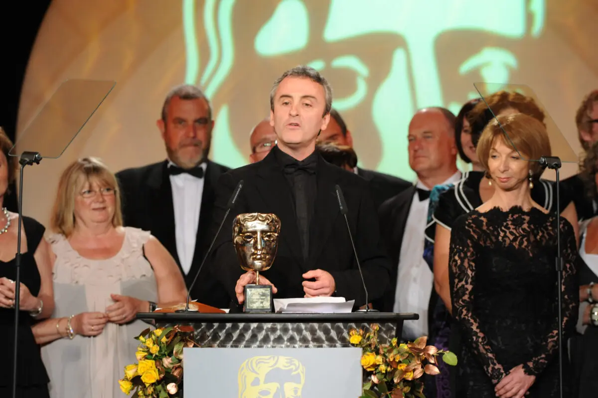 A man with grey hair wearing a black shirt and suit stands at a lectern with a gold BAFTA mask. He is surrounded by men in tuxedos and women in smart dresses.