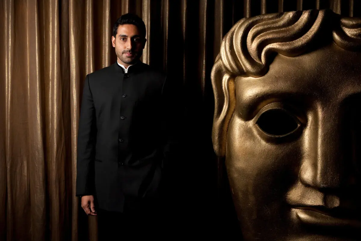 Abhishek Bachchan. A man with black hair and stubble wearing a black suit and stood next to a giant gold BAFTA mask.