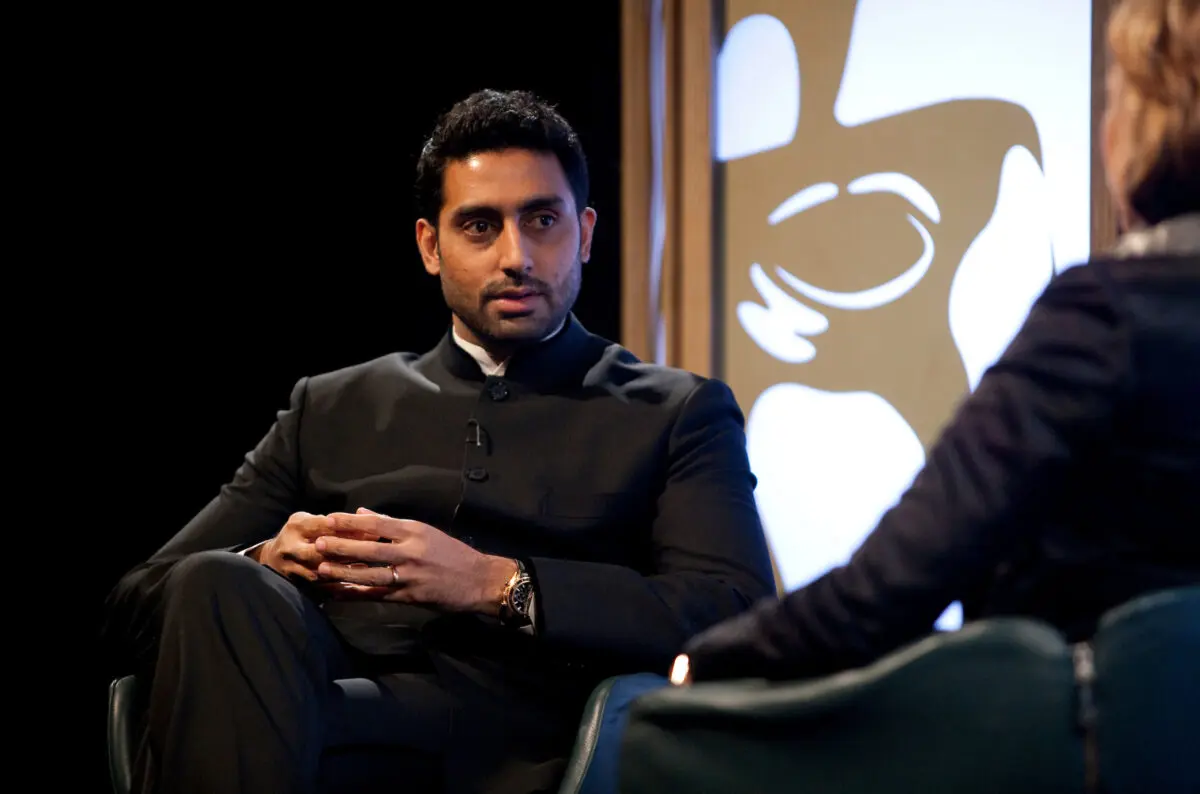 Abhishek Bachchan. A man with black hair and stubble wearing a black suit and sat on stage.