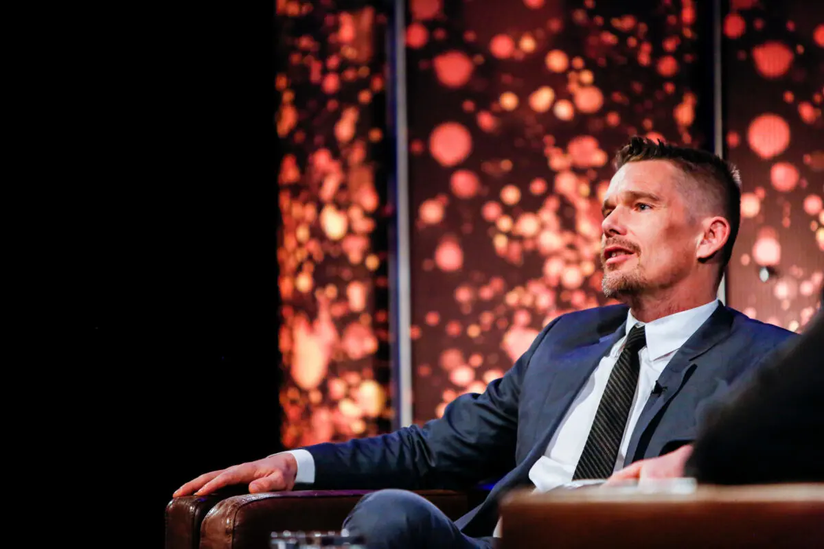A man with short cropped dark hair and a goatee sat in a brown leather chair with sparkly lights behind him. He is wearing a white shirt, grey suit and black tie.