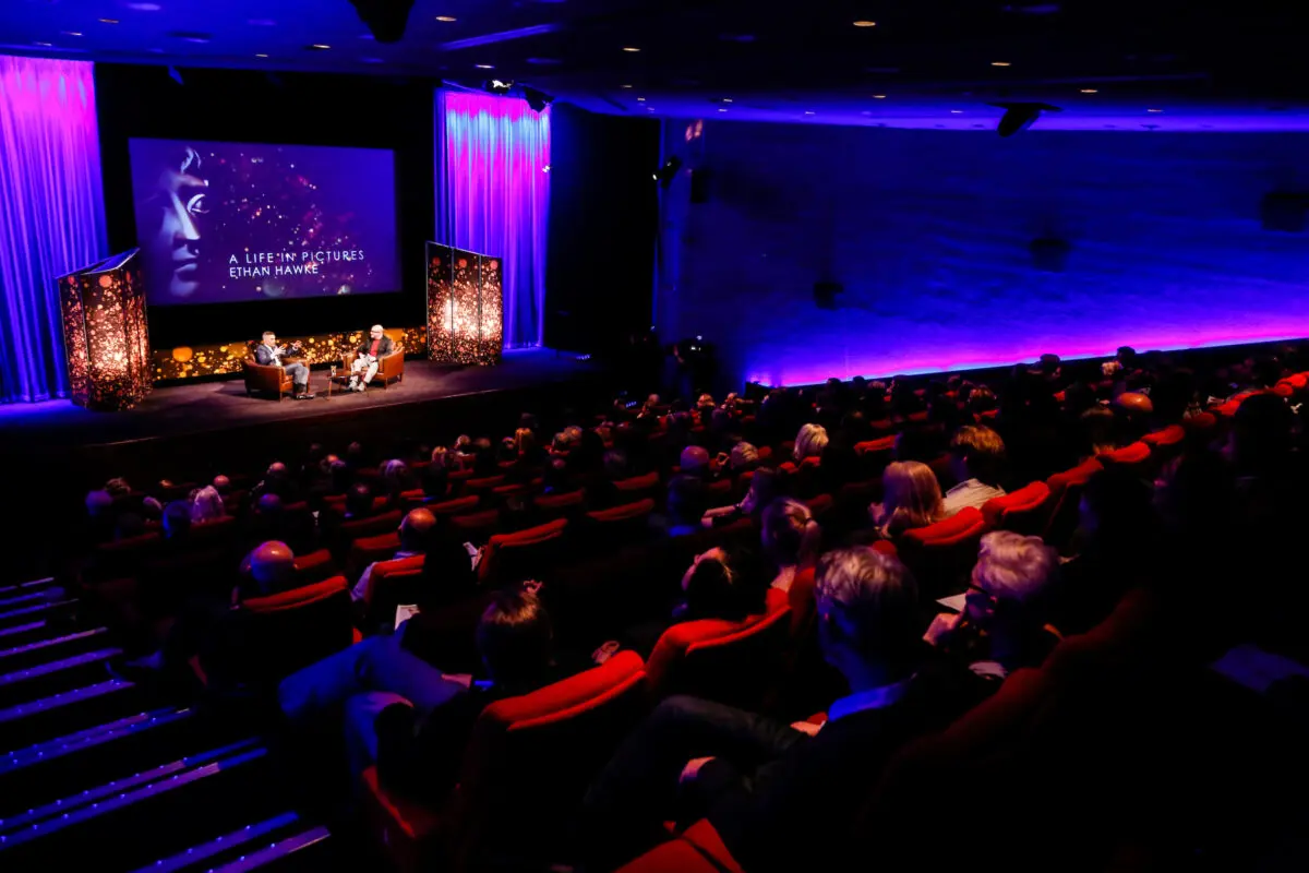 A cinema with red seats and low purple lighting photographed from above. All the chairs are facing towards a stage with two men sat in armchairs and a screen which reads 'A Life in Pictures: Ethan Hawke' behind them.