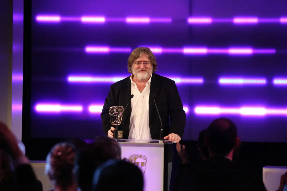 A white man with long brown hair, a white beard and small wire framed glasses. He is wearing a white shirt and black jacket and stood at a lectern with his hand on a BAFTA award.