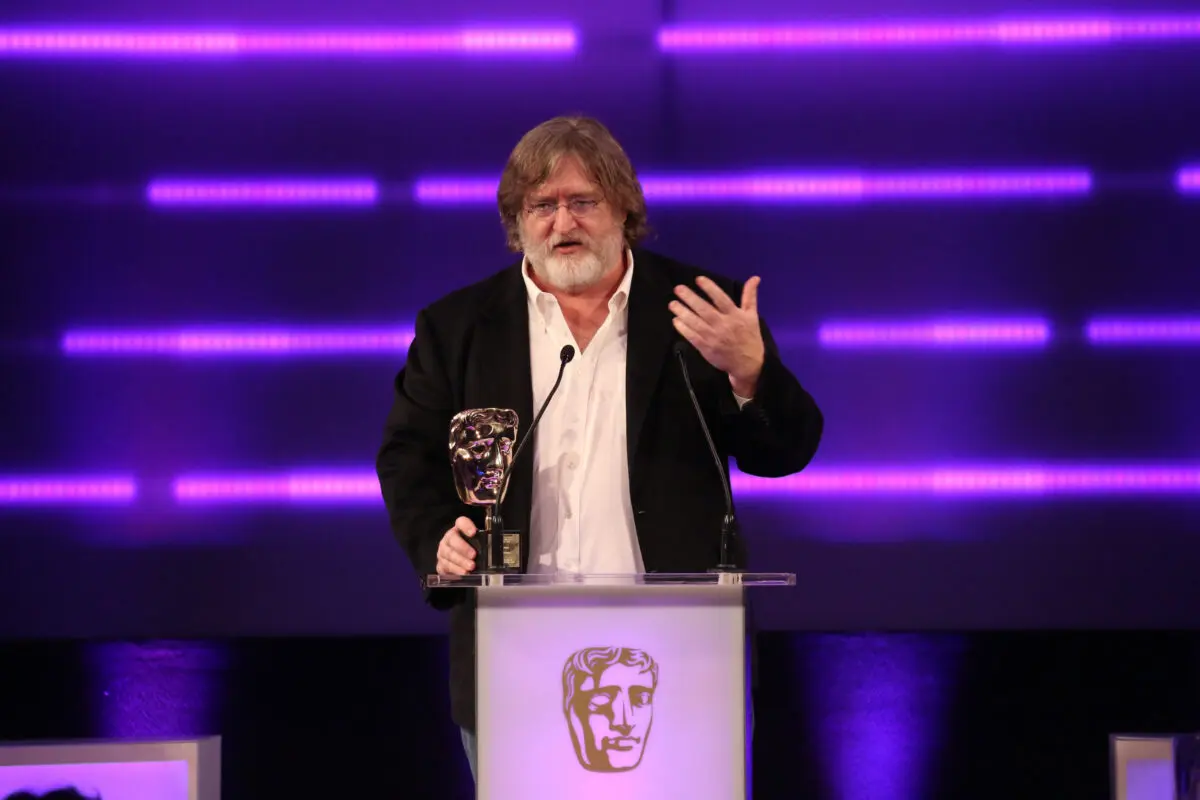 A white man with long brown hair, a white beard and small wire framed glasses. He is wearing a white shirt and black jacket and stood at a lectern with his hand on a BAFTA award.