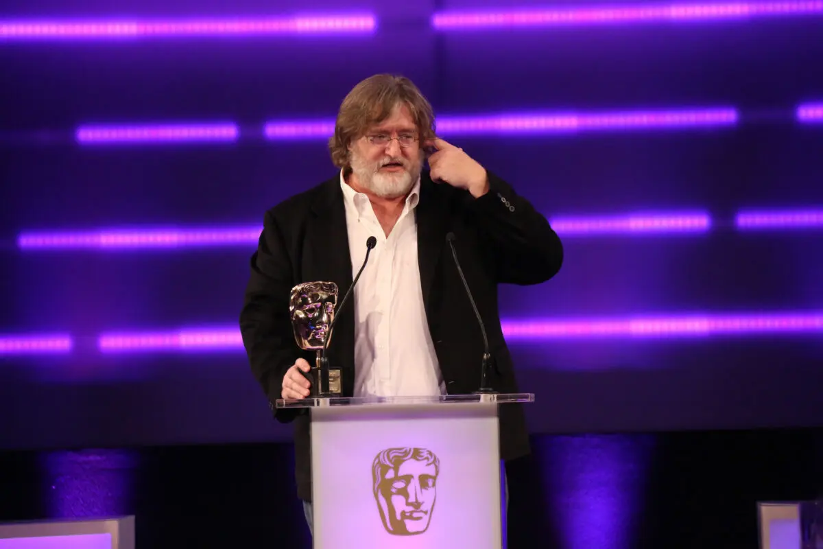 A white man with long brown hair, a white beard and small wire framed glasses. He is wearing a white shirt and black jacket and stood at a lectern with his hand on a BAFTA award.