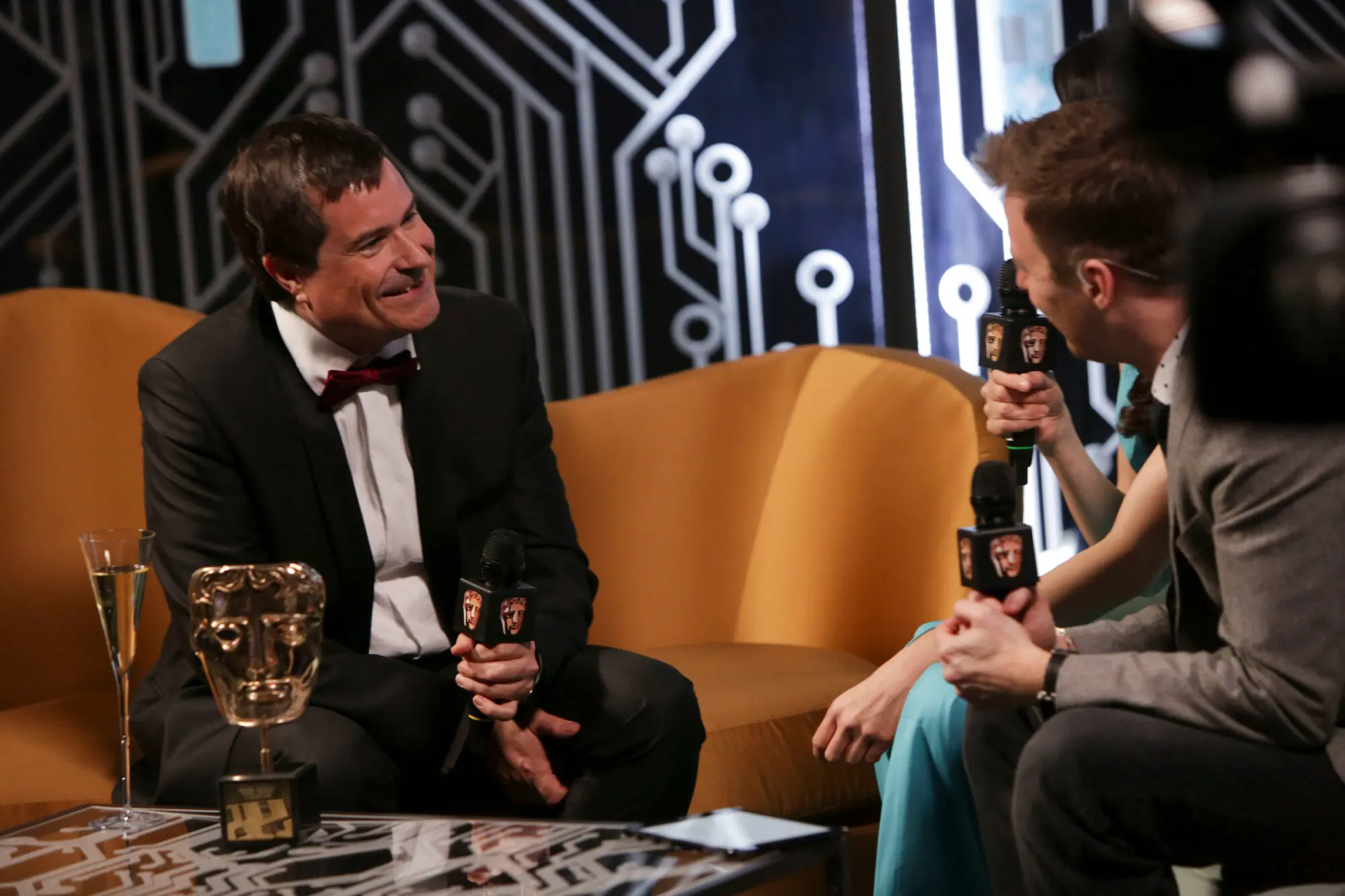 A white man with dark hair wearing a black suit and red bow tie sitting on an orange velvet sofa. He is holding a microphone and talking to a couple of interviewers who are sat opposite.