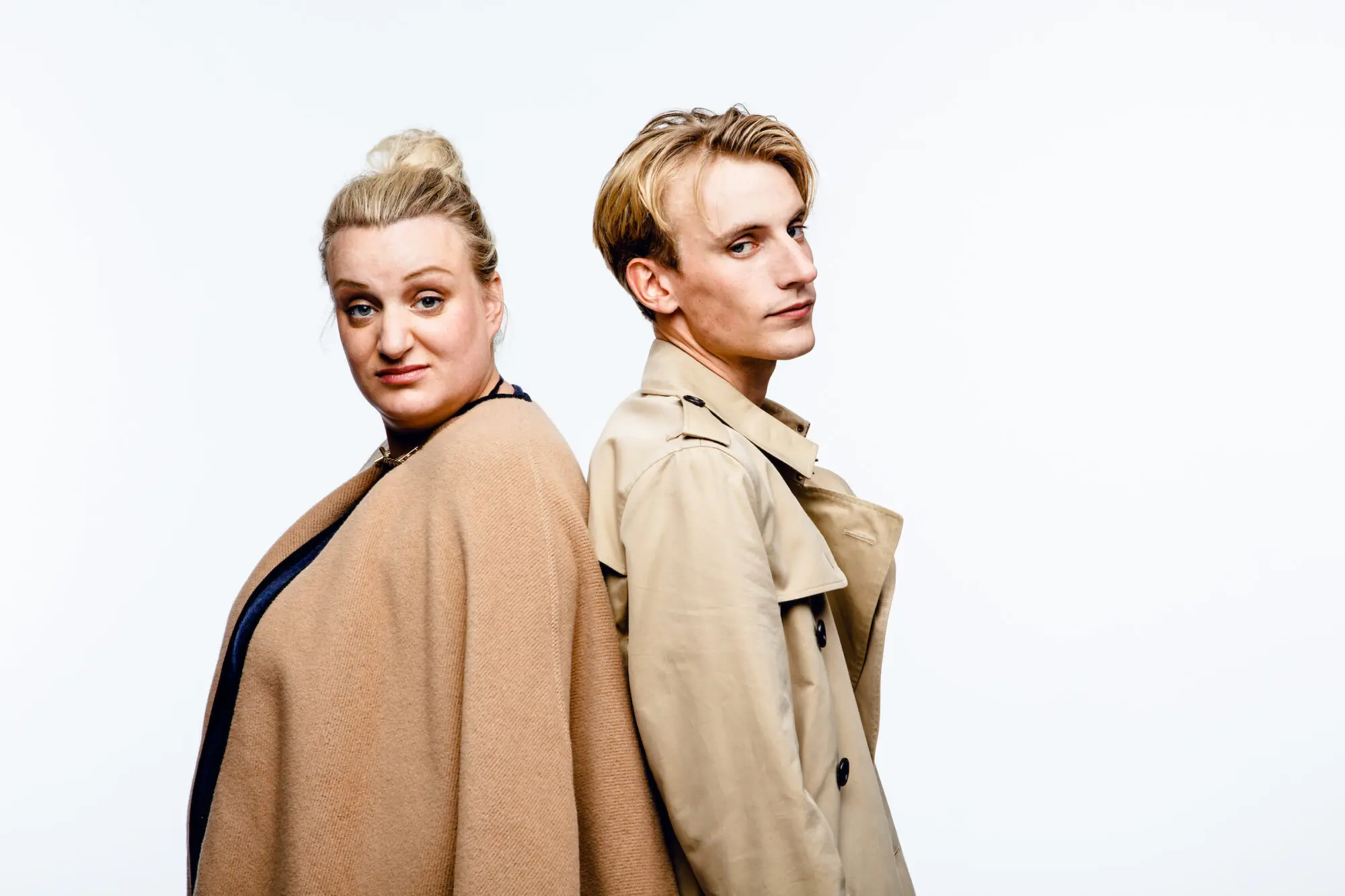 A blonde woman with hair tied in a bun and wearing a camel coloured cape, standing back to back with a blond man wearing a beige trench coat.