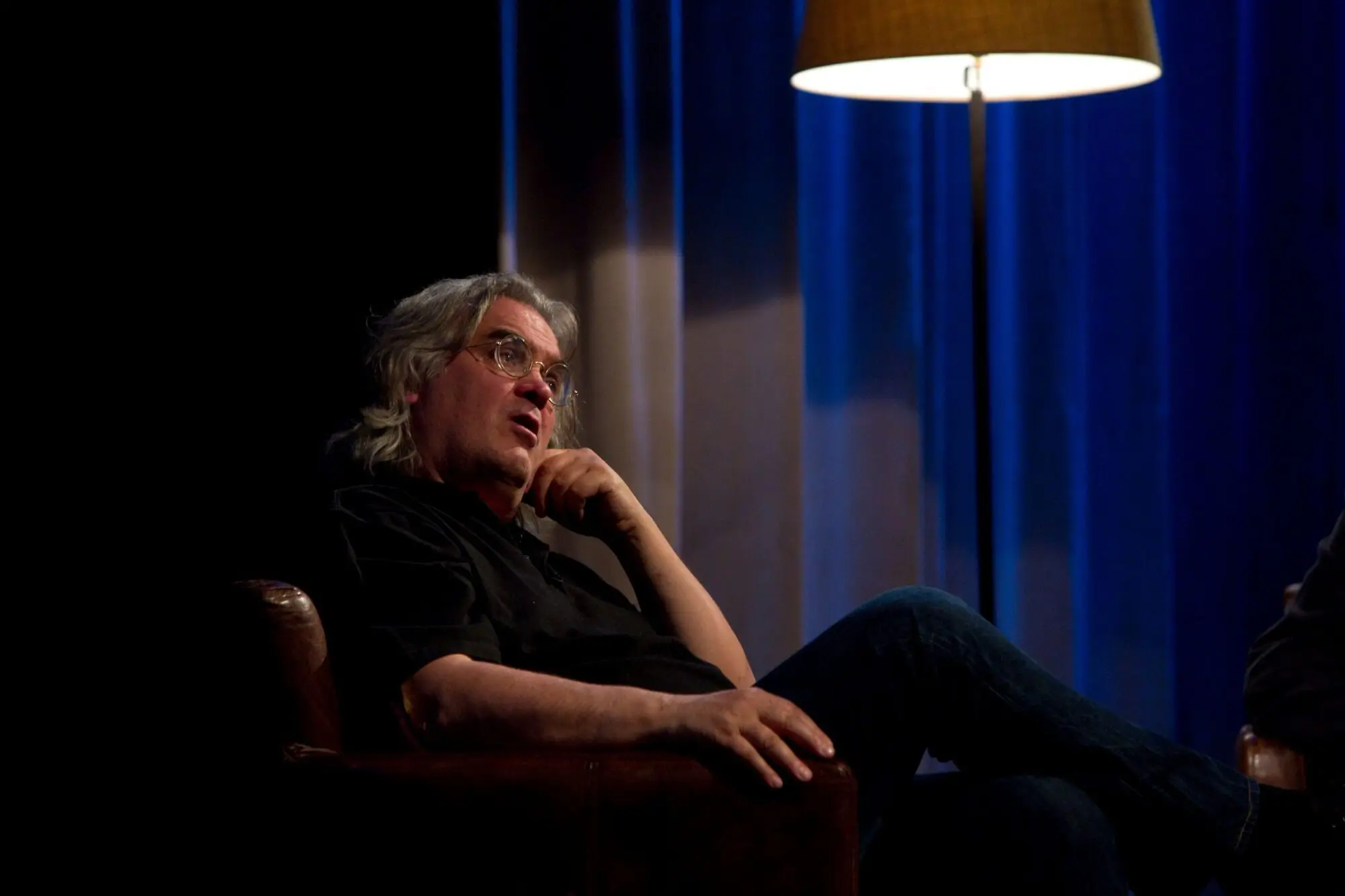 A man with long grey hair and glasses sat in a large armchair on a stage.