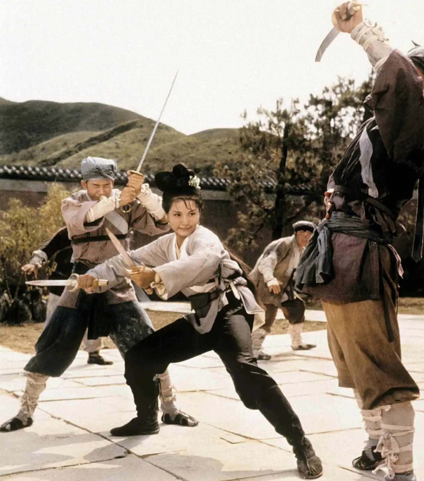An Asian woman with black hair tied up in a bun with a flower in it. She is in a squatting fighting stance and holding two knives preparing to do battle with two men that are holding swords and surrounding her.