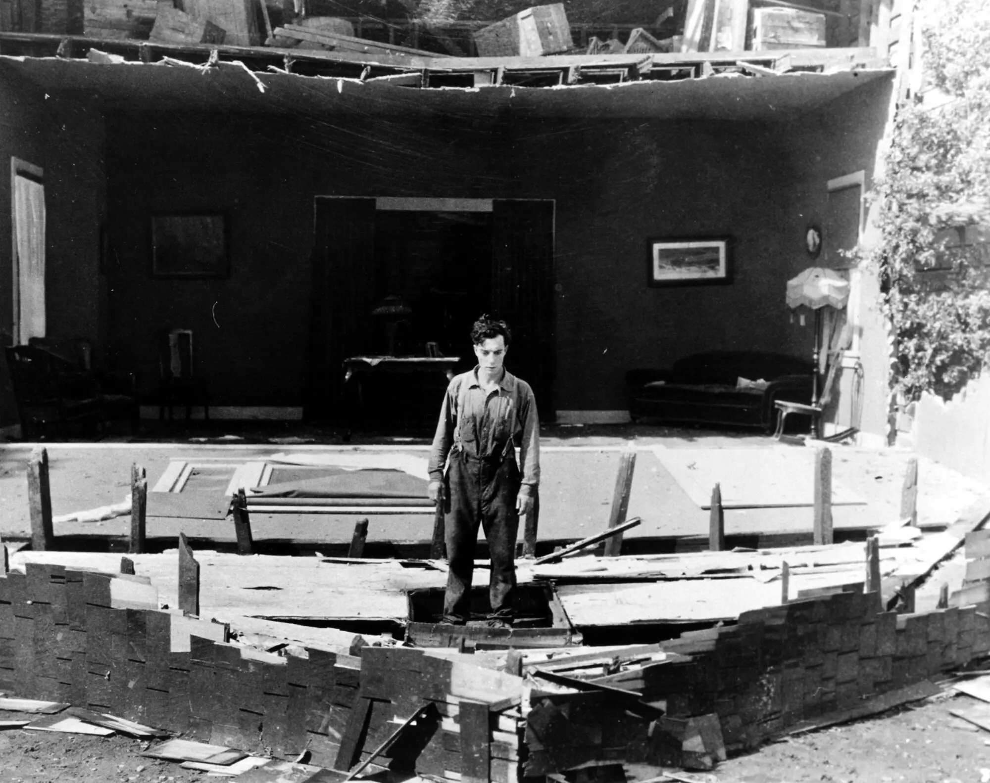A black and white photograph of a man stood in the remains of a house where the front has fallen off.