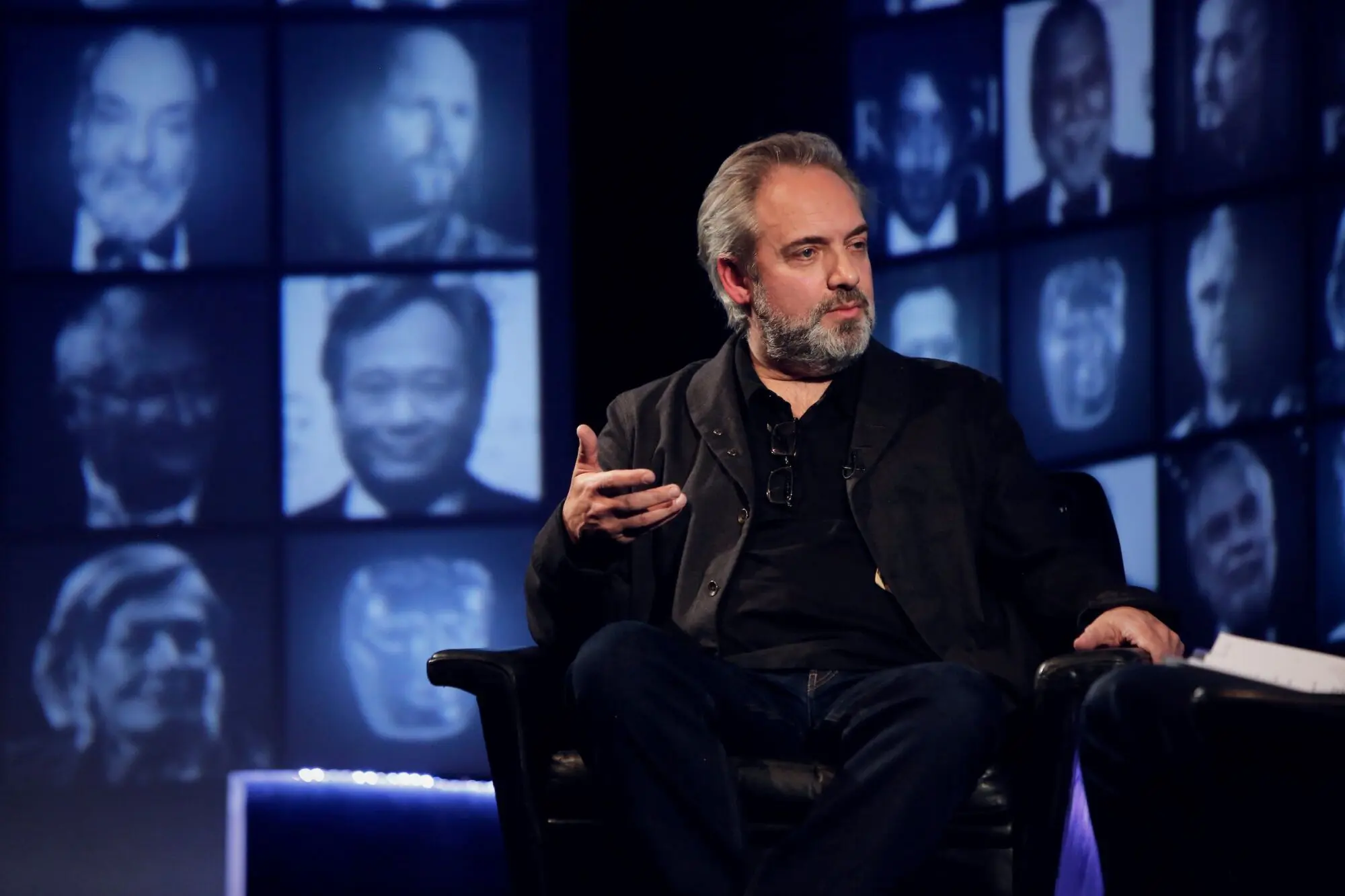 Sam Mendes. A white man with thick grey hair wearing a black shirt and suit.