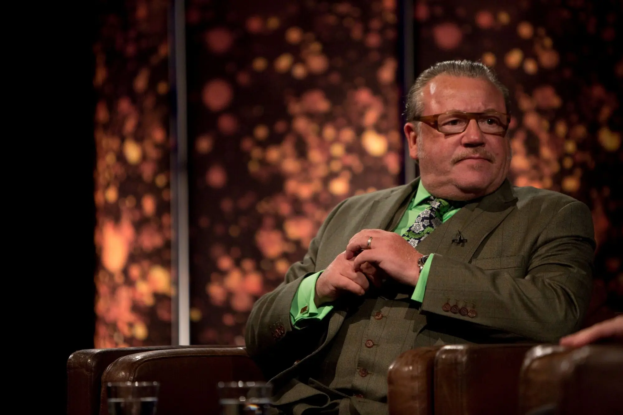 Ray Winstone. A white man with slicked back grey hair wearing rectangular acetate glasses. He is wearing a dark green tweed waistcoat and suit with a bright green shirt and floral tie.