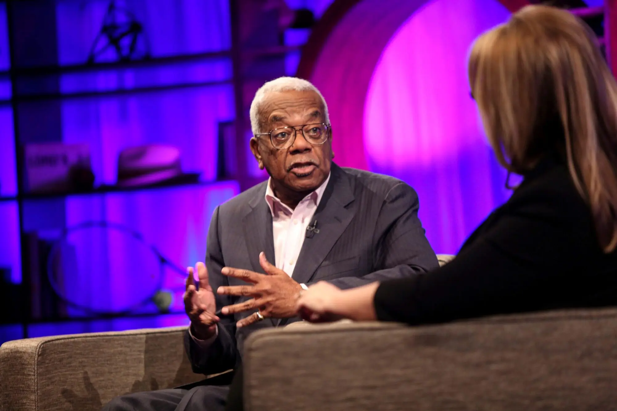 A black man with white hair and wire framed aviator style glasses. He is wearing a white shirt and grey suit jacket.