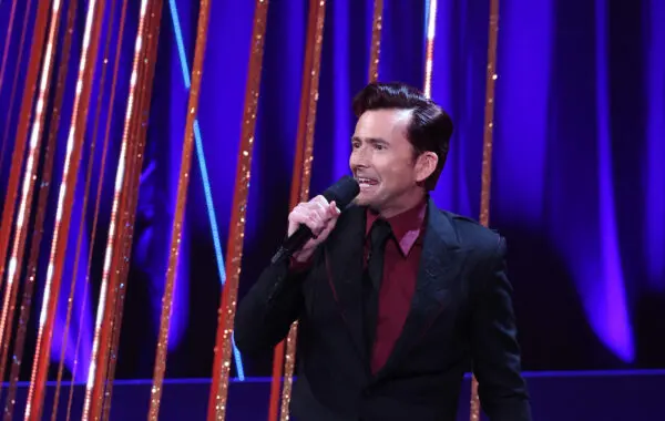 David Tennant. A man with thick dark hair combed to one side. He is wearing a dark red shirt, black suit jacket with embossed lapels, black tie, and is holding a microphone.