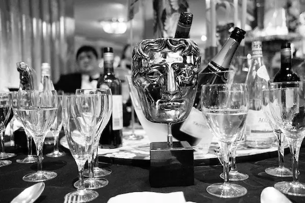 Black and white image of a BAFTA mask on a table surrounded by wine glasses.