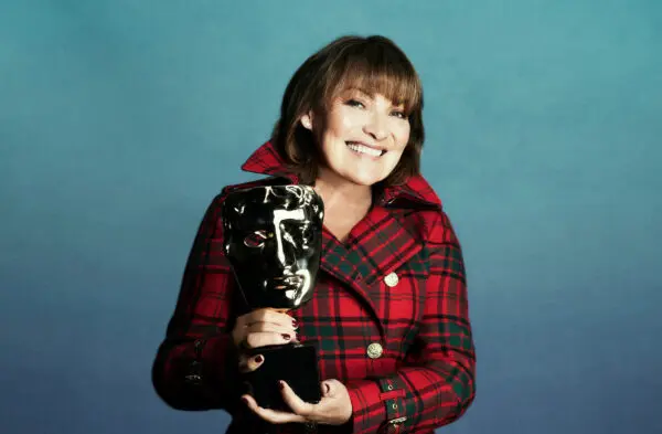 Lorraine Kelly. A white woman with dark bobbed hair and a thick fringe. She is wearing a tartan double breasted jacket with upturned collar and is holding a BAFTA award.