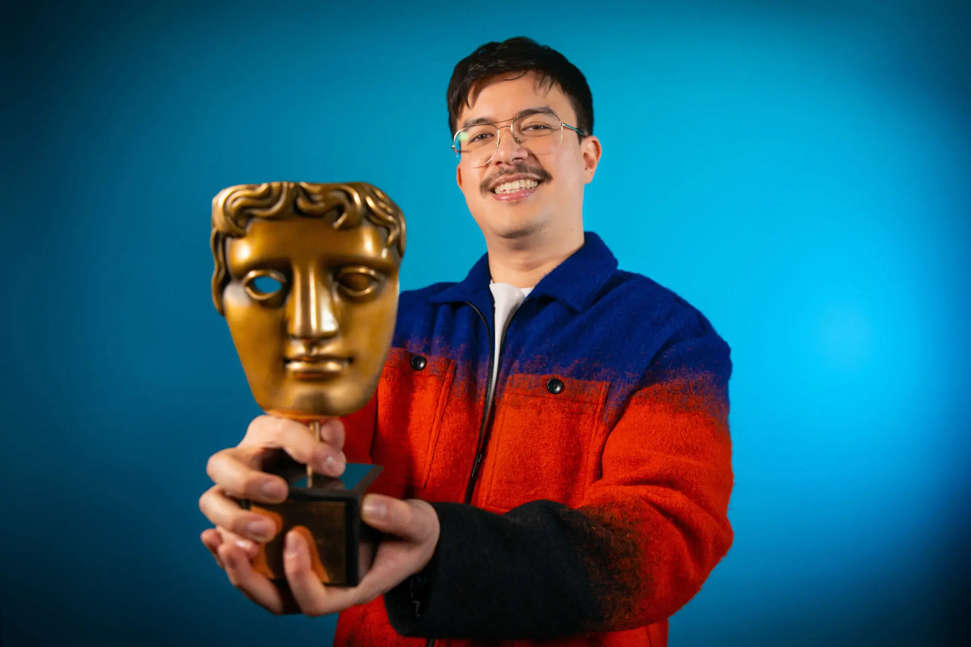 Phil Wang. An Asian man with wire framed glasses and wearing a blue and red zip up top, holding a BAFTA award.
