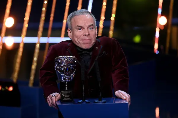 A white man with grey hair is stood in front of a podium with a BAFTA Award on it.