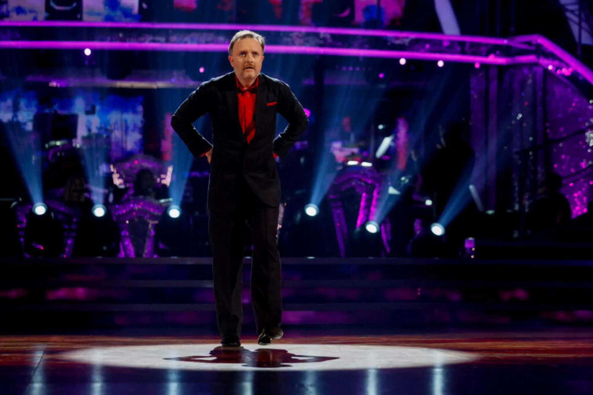 A white man with grey hair, wearing a red shirt and black suit, stood in the middle of a dancefloor surrounded by spotlights and purple tube tights. 