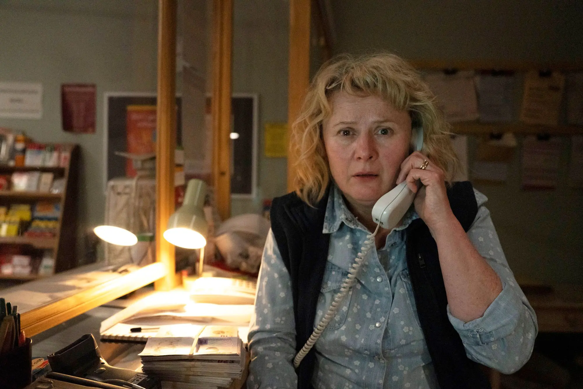 A close up of a white woman with short curly blonde hair on the phone. She is surrounded by a desk and papers and looks like she is in shock.