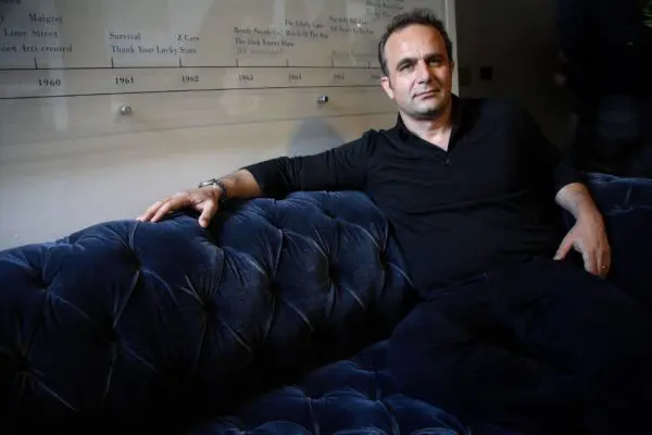 Pietro Scalia. A white man with short dark hair wearing a black shirt with an unbuttoned collar. He is sat on a dark blue velvet sofa with one arm draped over the back of it.