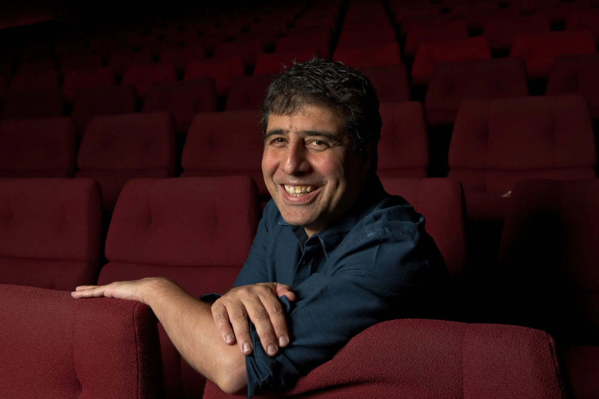Hossein Amini. An Iranian man with thick, short dark hair wearing a blue shirt and sat in a red cinema seat.