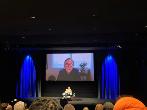 A man sat on a chair in the middle of a stage and another man projected on a large screen behind him.