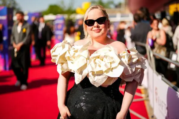 Nicola Coughlan, a white blonde woman stood on a red carpet wearing a full length black dress with white rose collar and statement black sunglasses.