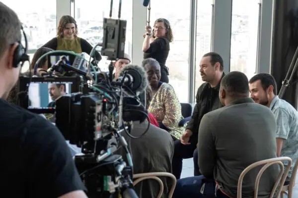 A white man in a dark suit being interviewed by a black lady in a gold sparkly jacket while camera crews stand around them.