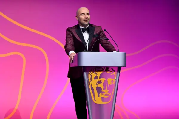 Lee Walters. A man wearing a pink jacket, white shirt and black bow tie stands at a silver podium adorned with a gold BAFTA mask logo.