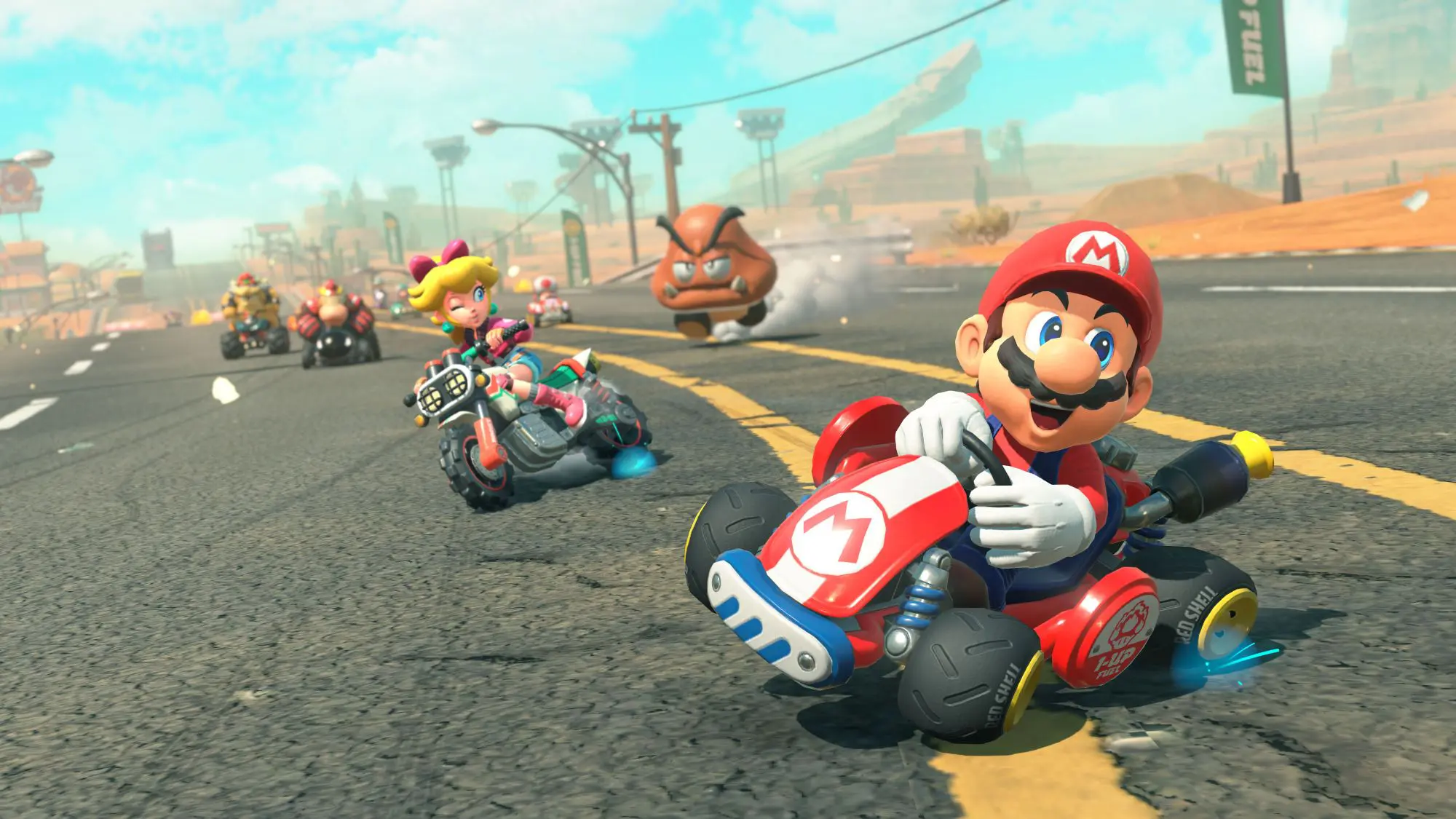 A still from the game Mario Kart World. Five racers are seen on a race track speeding away from what looks like a brown blob with a face. Mario is at the front wearing his all red and blue outfit and 'M' cap.