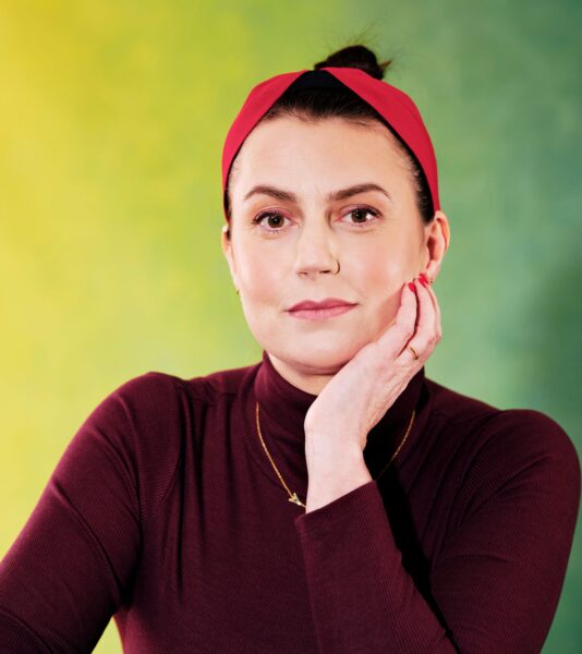 Jemma Gander. A woman with dark hair tied back in a high bun and with a red hairband on. She is wearing a burgundy polo-neck and short gold chain necklace and is leaning her face on her hand.