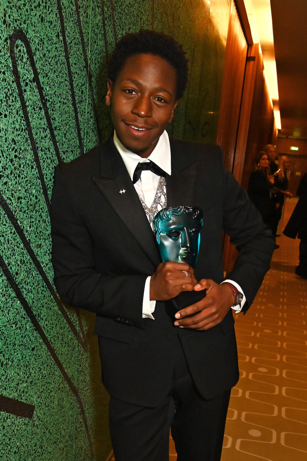 David Jonsson. A young black man with thick dark hair wearing a black tuxedo, white shirt and silver open knit vest and holding a blue EE Rising Star BAFTA.