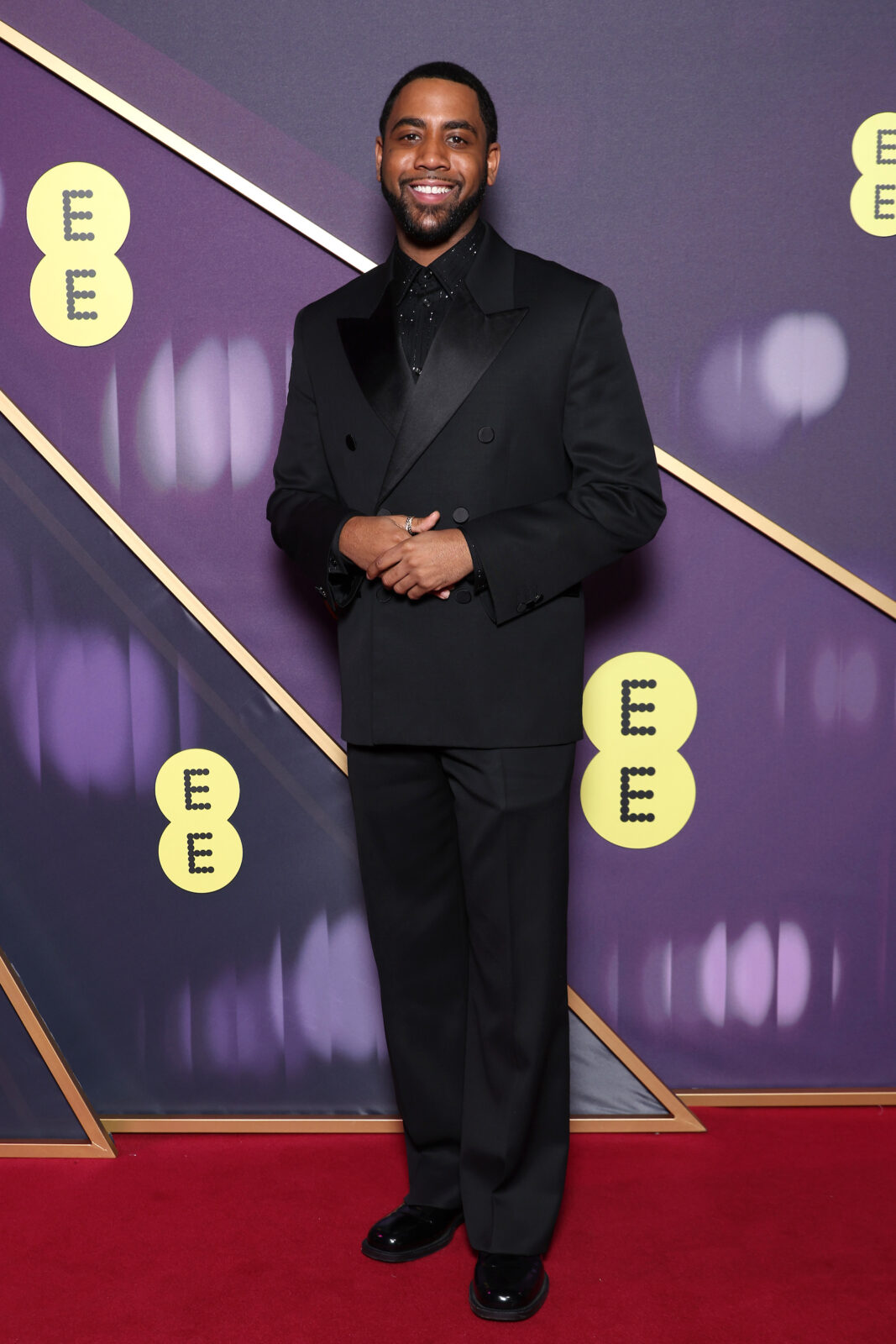 Jharrel Jerome. A black man with short cropped dark hair and trimmed facial hair. He is wearing a black suit with black shirt underneath.