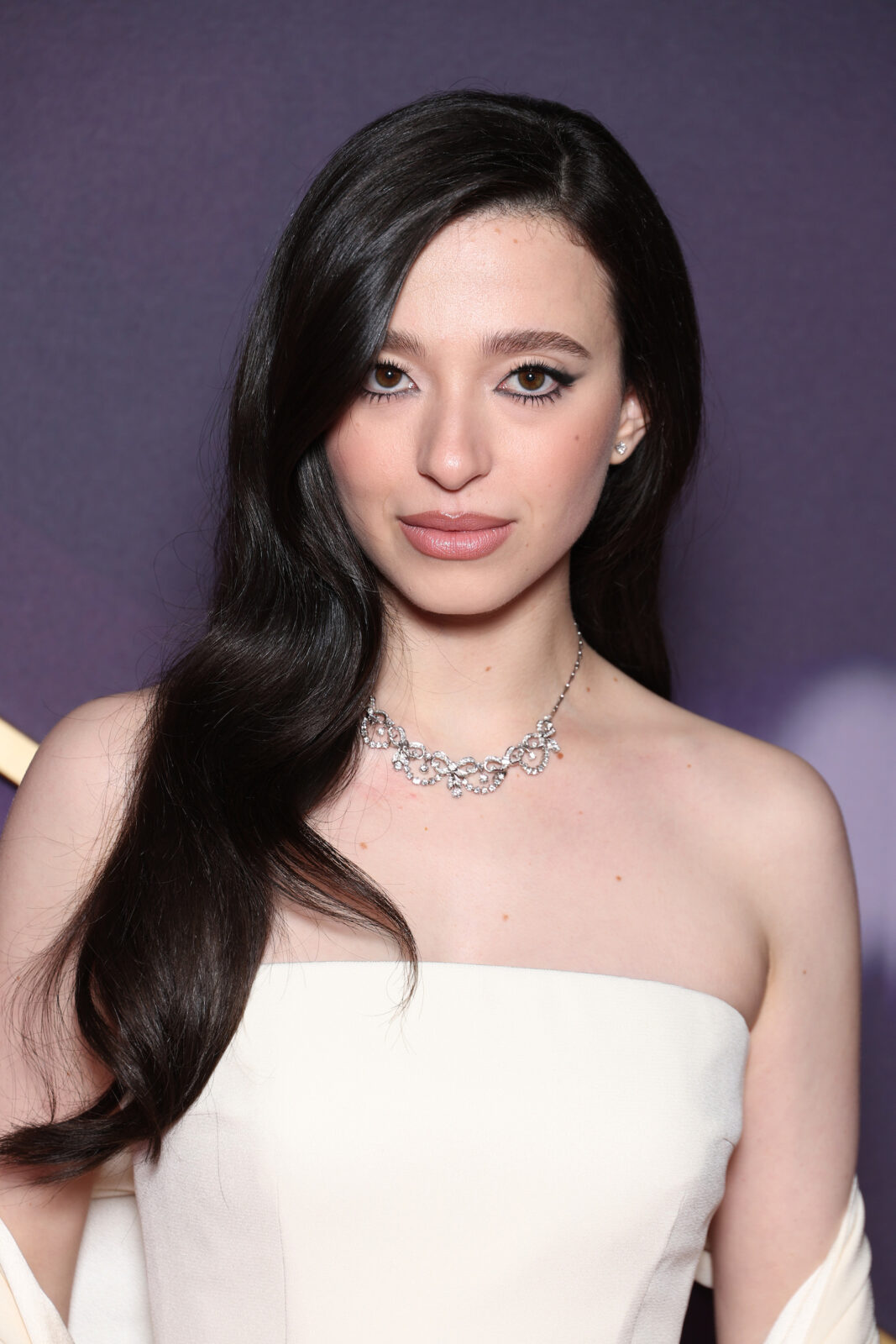 Mikey Madison. A woman with long dark hair wearing a white strapless evening gown and white scarf.