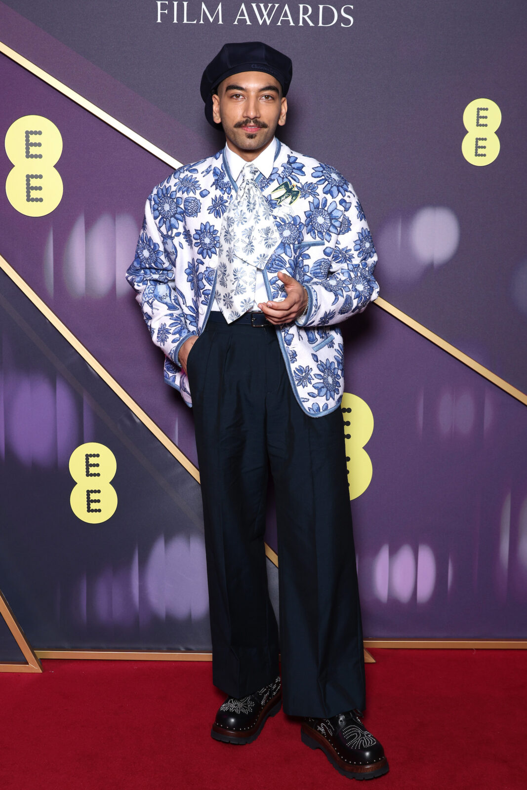 Nabhaan Rizwan. A man with a dark moustache wearing high waisted dark trousers, blue and white floral patterned jacket, patterned tie and a black hat.