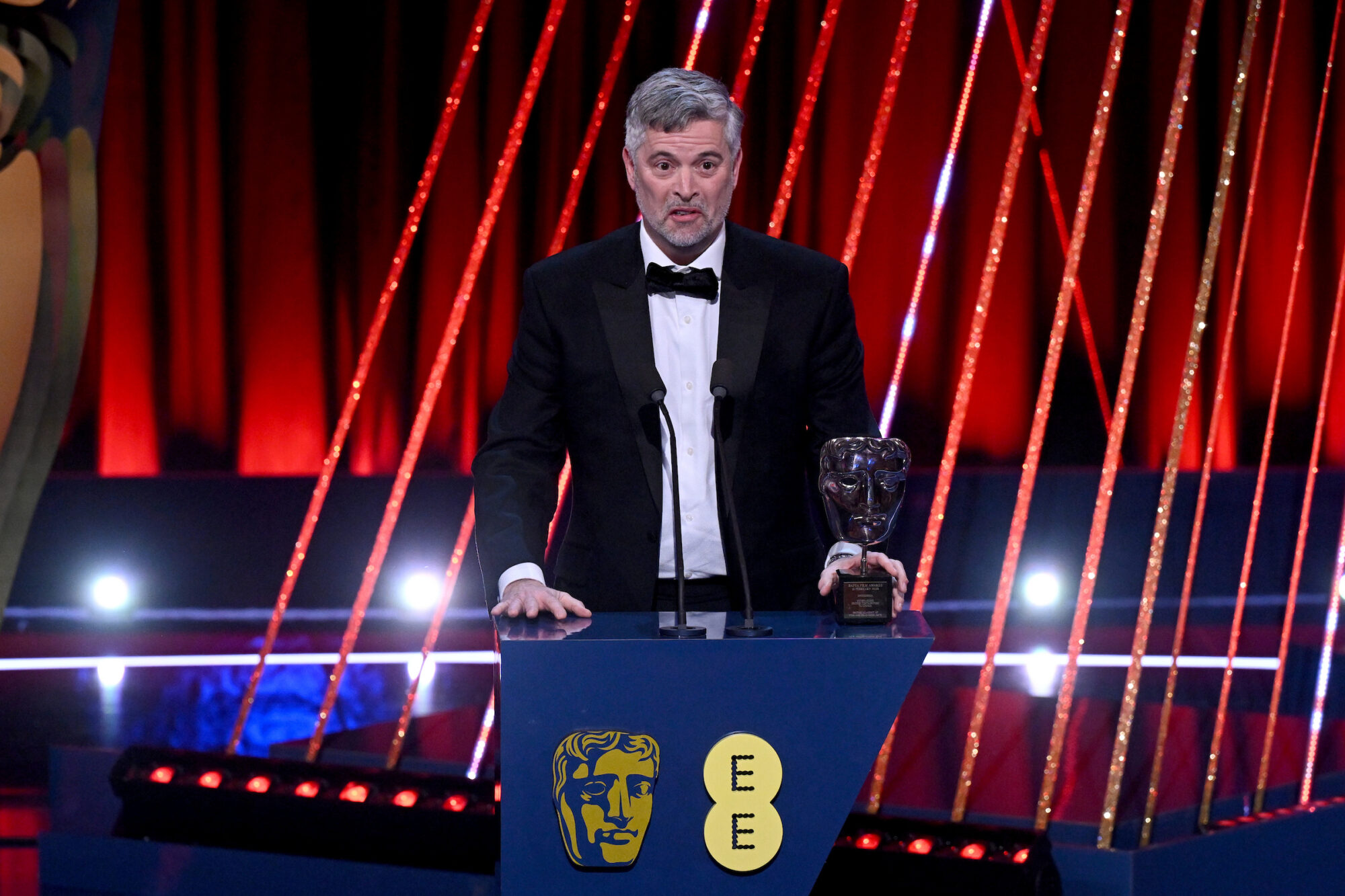 Colin Lawrence accepts the Outstanding British Contribution to Cinema Award for MediCinema. A man with thick grey hair wearing a tuxedo and bow tie standing at a podium on stage.