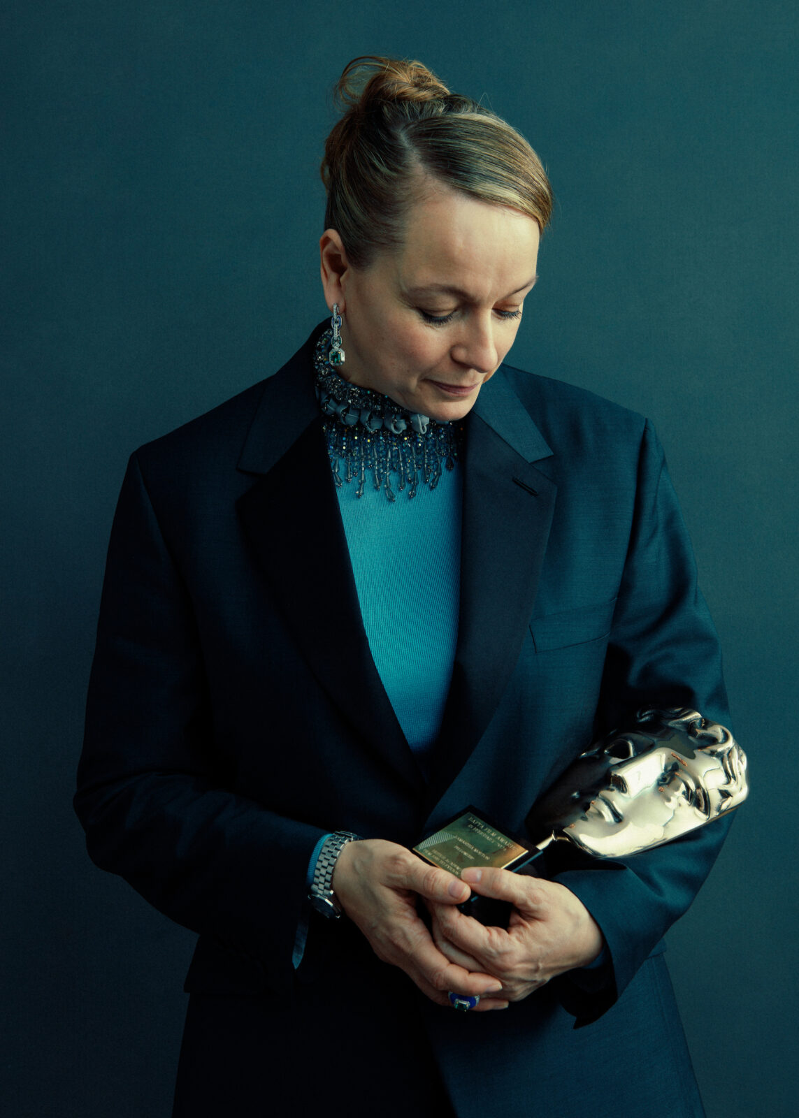 Samantha Morton. A white woman with blonde hair tied back in a bun. She is wearing a dark blue suit jacket over a turquoise top and is cradling a BAFTA in her arms.