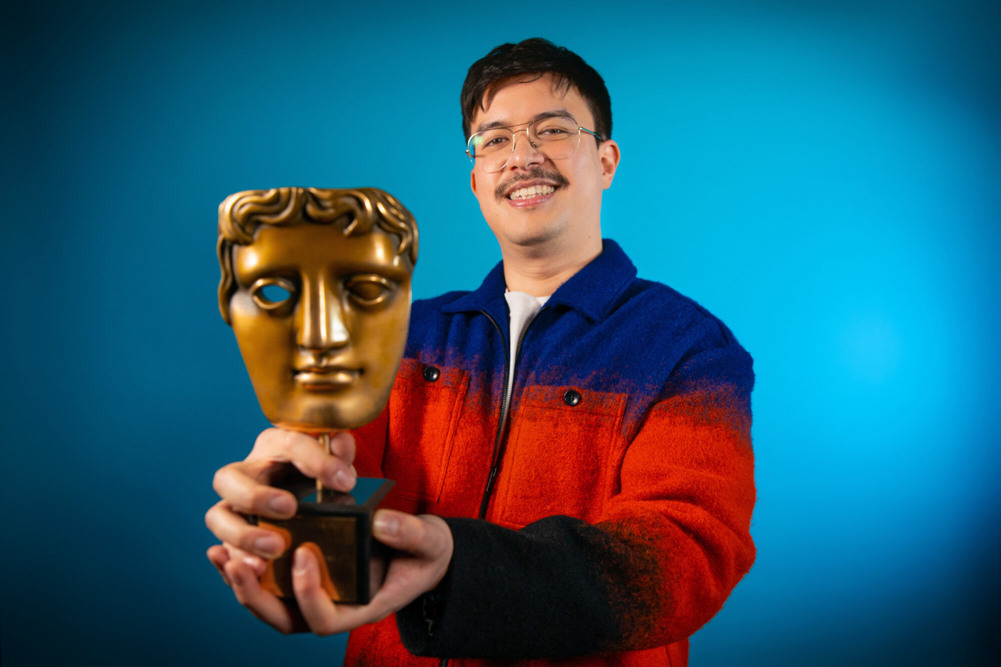 Phil Wang. An Asian man with wire framed glasses and wearing a blue and red zip up top, holding a BAFTA award.